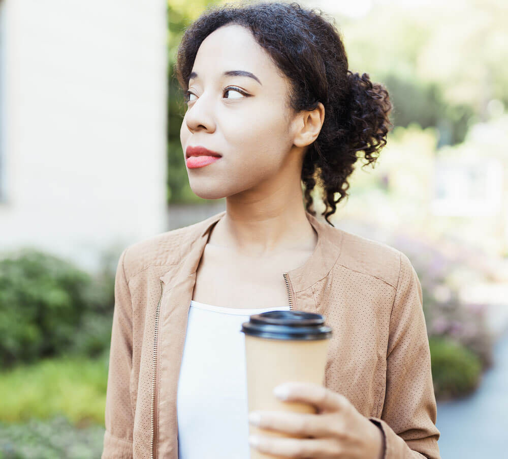Young African multiracial woman holding disposable brown paper coffee cup on street outdoors